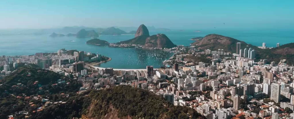 Rio de Janeiro (RJ) Visão aérea de vários prédios cercados pelo mar no litoral com o morro do Pão de Açúcar ao fundo.