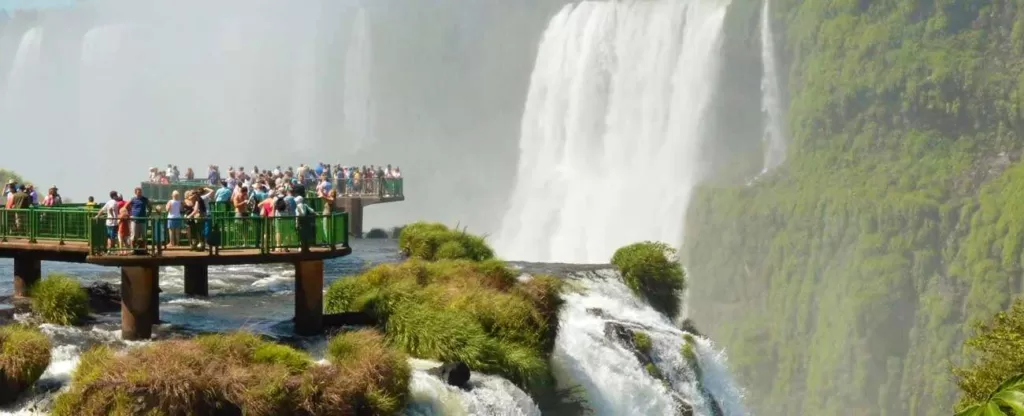Passarela sobre um rio com várias pessoas observando uma cachoeira ao longe.