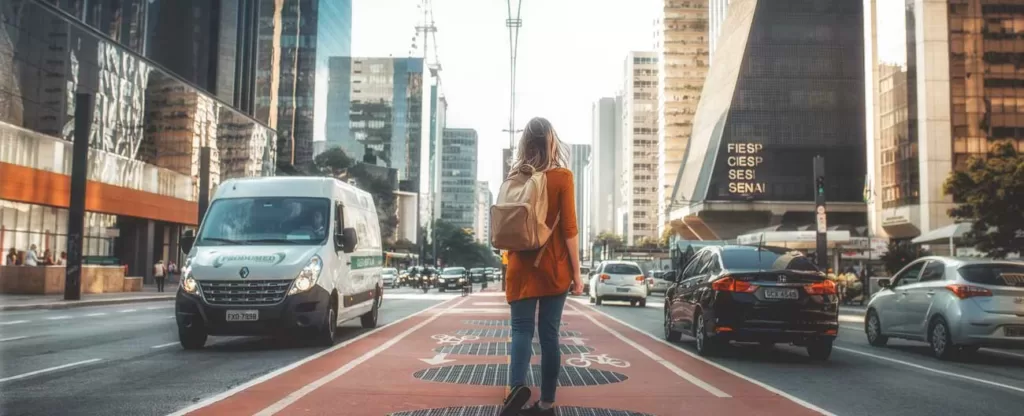 Mulher com mochila nas costas andando no meio da movimentada Avenida Paulista com trânsito e prédios altos dos dois lados.
